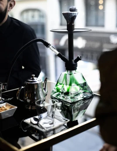 Une chicha en verre vert posée sur une table en verre noir réfléchissante, avec une main qui verse de l'eau à l'aide d'une théière en métal.