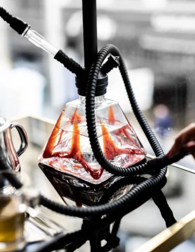 Une chicha moderne avec un design transparent et des tuyaux noirs, posée sur une table en verre avec des reflets lumineux.
